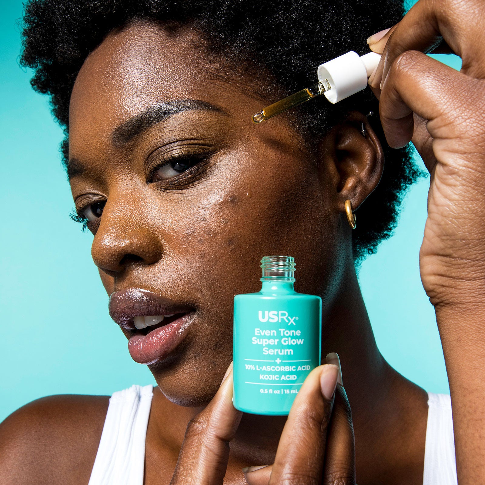Woman applying Urban Skin Rx skincare Even Tone Super Glow Serum to her face with a dropper against a teal background