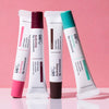 Multiple tubes of USRx Hydrating Lip Treatment in various tinted shades on a pink background